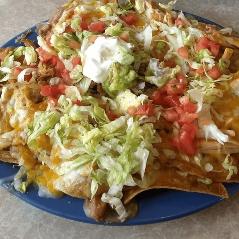 A large plate of Carmela's Mexican Restaurant nachos, topped with cheese, lettuce, tomatoes, sour cream, and guacamole.