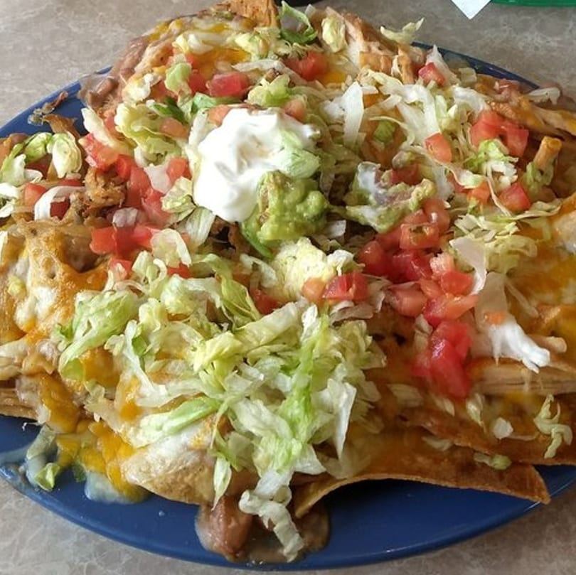 A large plate of Carmela's Mexican Restaurant nachos, topped with cheese, lettuce, tomatoes, guacamole, and sour cream.