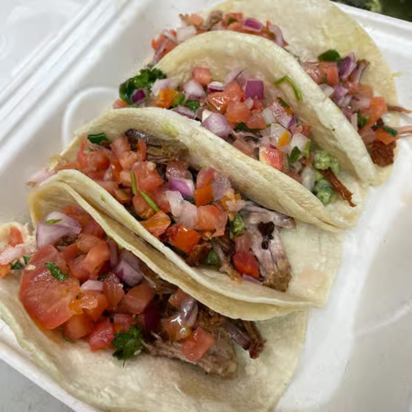 Three Carmela's Mexican Restaurant tacos filled with meat, pico de gallo, and guacamole in a white container.